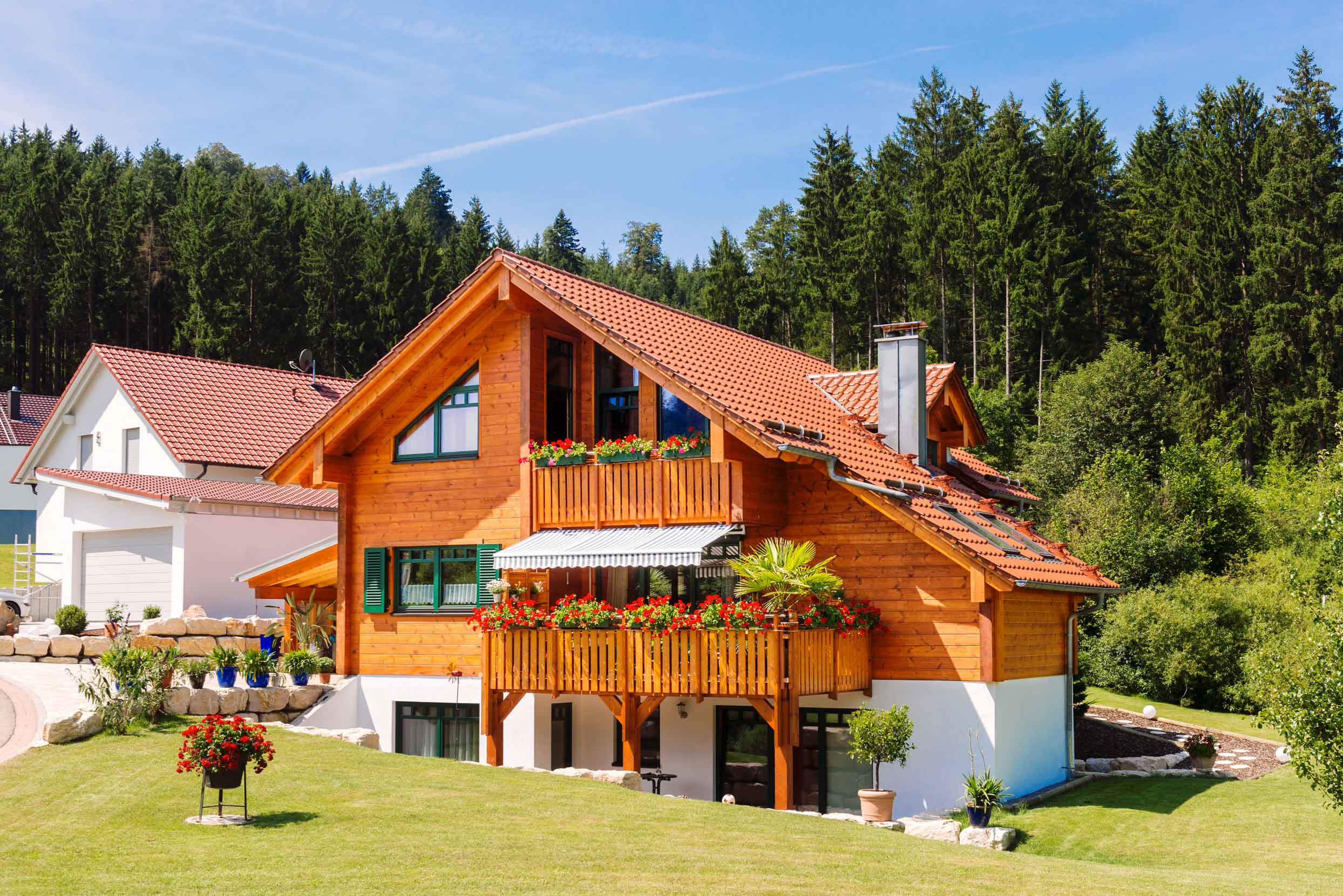 Holzhaus im Landhaus-Stil mit Kiefernfassade, tannengrünen Fensterläden und zweigeschossigen Holzbalkonen vor bewaldeter Hanglage